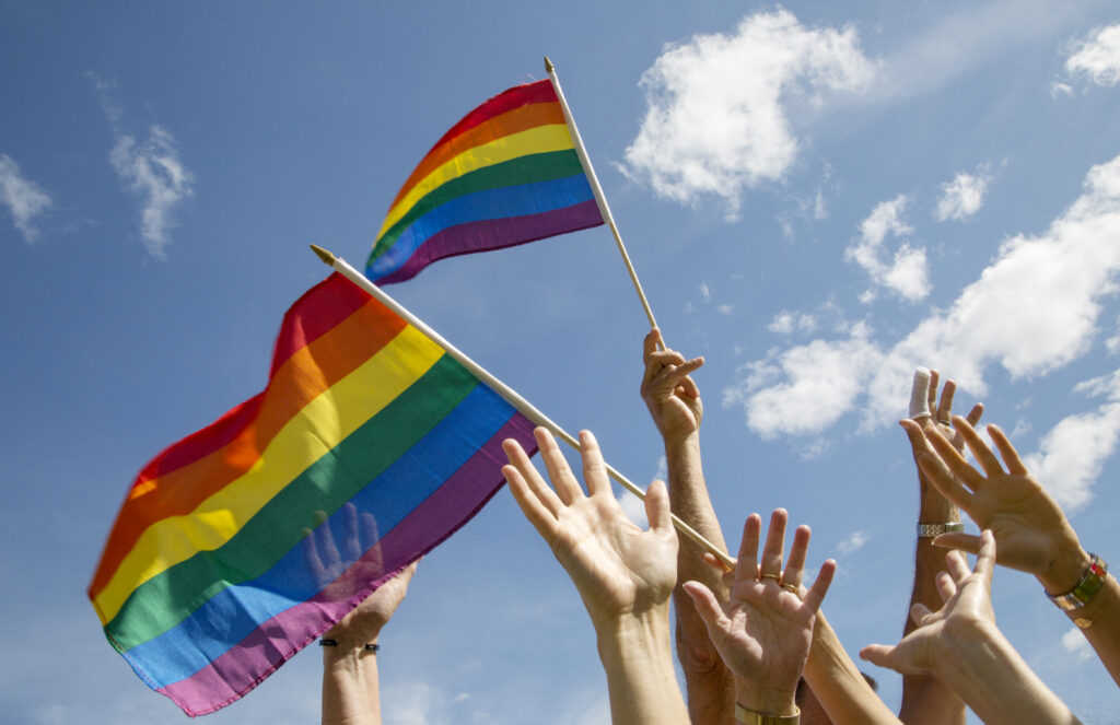 Flera händer sträcks upp mot en blå himmel. Händerna håller i två regnbågsflaggor.